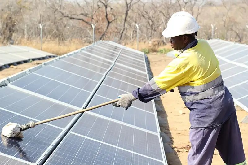 Worker in safety gear cleaning a row of ground-mounted solar panels with a long brush